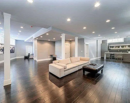 Open gray finished basement with white sofa, dark wood floor, columns, recessed lighting, and bar area