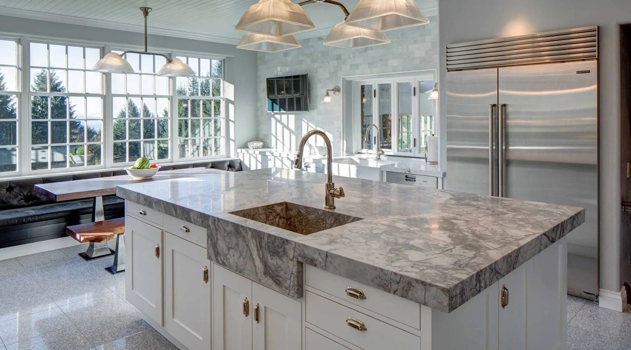 Bright kitchen with marble waterfall island, white cabinetry, large windows, and stainless refrigerator