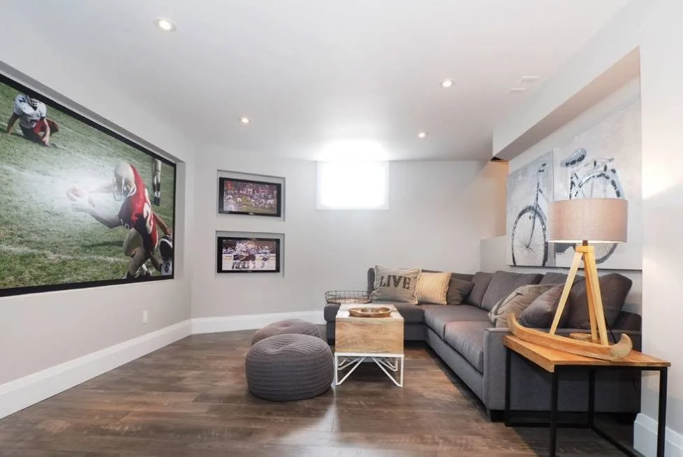 Basement sofa media room with large sports screen, gray sectional, ottoman, and recessed lights